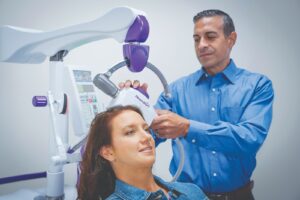 A male doctor adjusting a NeuroStar medical device positioned against the head of a smiling female patient during a treatment session.