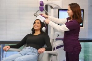 Technician in purple scrubs positions a TMS device on a smiling patient's head during treatment.
