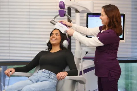 Technician in purple scrubs positions a TMS device on a smiling patient's head during treatment.